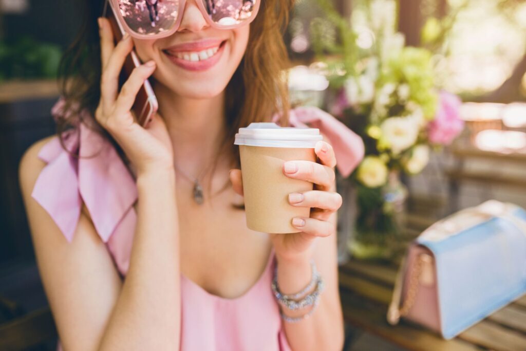 Portrait young attractive woman sitting cafe summer fashion outfit pink cotton dress sunglasses smiling drinking coffee stylish accessories trendy apparel talking phone