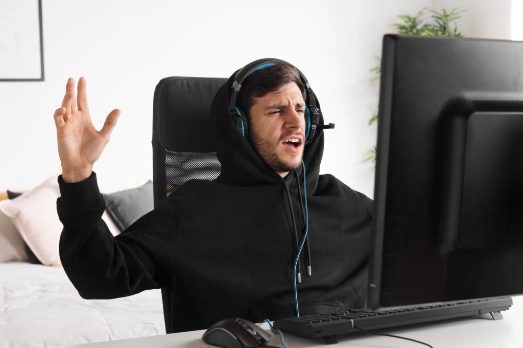 一名戴著耳機的男性玩家坐在電腦前,雙手舉起神情激動,疑似在進行線上對戰遊戲時情緒失控。